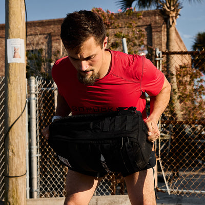 A man wearing a red Men’s Performance Tee - ToughMesh by GORUCK exercises outdoors, lifting a weighted backpack near a chain-link fence. The quick-drying capabilities of his gear ensure comfort under the sun, while a brick building and palm trees in the background cast shadows on his focused expression.