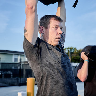 A person in a gray GORUCK Men’s Performance Tee - ToughMesh lifts a sandbag overhead, appearing focused in their outdoor workout. Another person is partially visible against the backdrop of a fenced area and the clear blue sky of the Italian Alps.