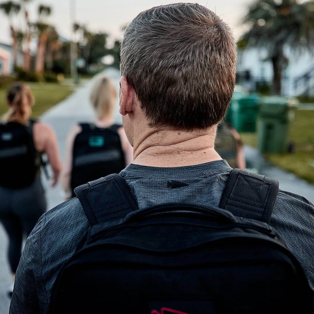 A group of people, viewed from behind, are walking up a paved road in a suburban setting with trees and houses along the street. They are dressed in casual attire, featuring backpacks and GORUCK's Men’s Performance Tee - ToughMesh, which is ideal for leisurely suburban strolls.