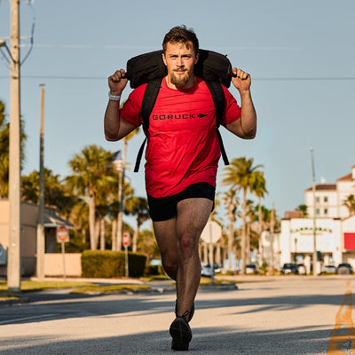 A man wearing a red Men’s Performance Tee featuring GORUCK ToughMesh fabric jogs down a sunny street, his black backpack securely fastened. The lightweight training tee fits comfortably as palm trees border the road and buildings rise in the background.