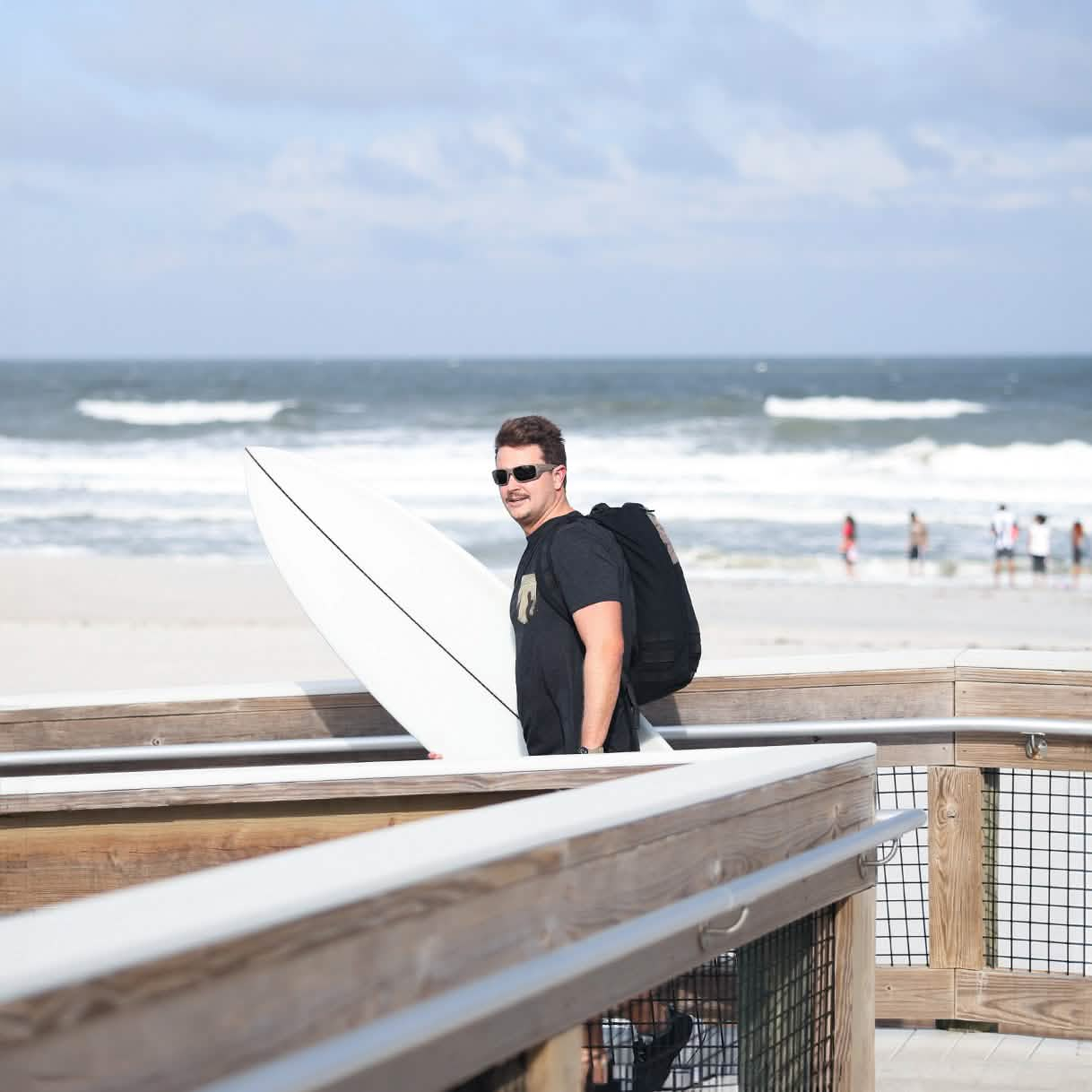 A person with a mustache and sunglasses stands on a boardwalk by the beach, holding a white surfboard and wearing a black backpack. Sporting the GORUCK x Birdwell Tee - Tri-Blend, they are surrounded by ocean waves crashing in the background, while a few people stroll along the sandy shore.