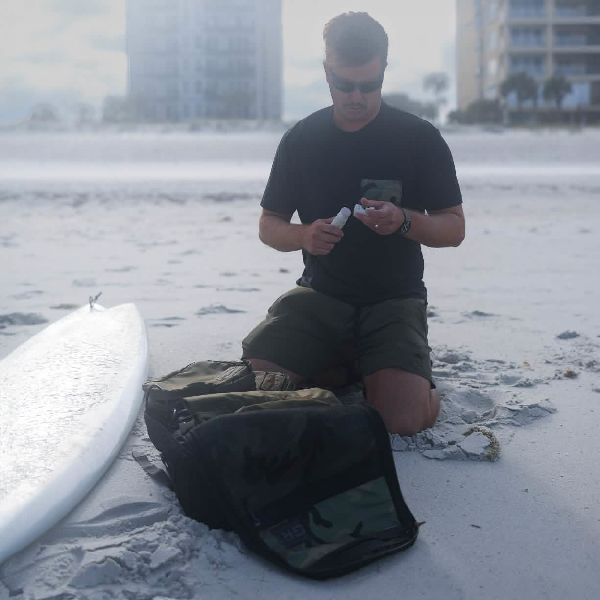 Kneeling on a sandy beach, a person wearing a GORUCK x Birdwell Tee - Tri-Blend and green shorts applies sunscreen next to their surfboard. An open camouflage-patterned backpack, famed for its GORUCK Scars durability, rests nearby. In the background, high-rise buildings and palm trees punctuate the skyline.