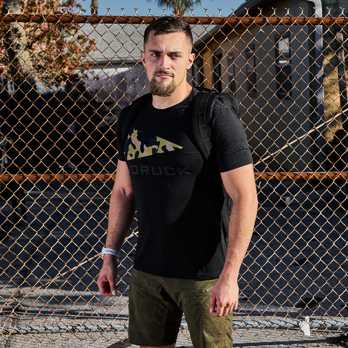 A man with a beard stands in front of a chain-link fence, wearing a premium black GORUCK Spearhead Tee from vendor-unknown and green shorts. He carries a black GORUCK Spearhead backpack and has a bracelet on his wrist. The setting appears to be outdoors on a sunny day.