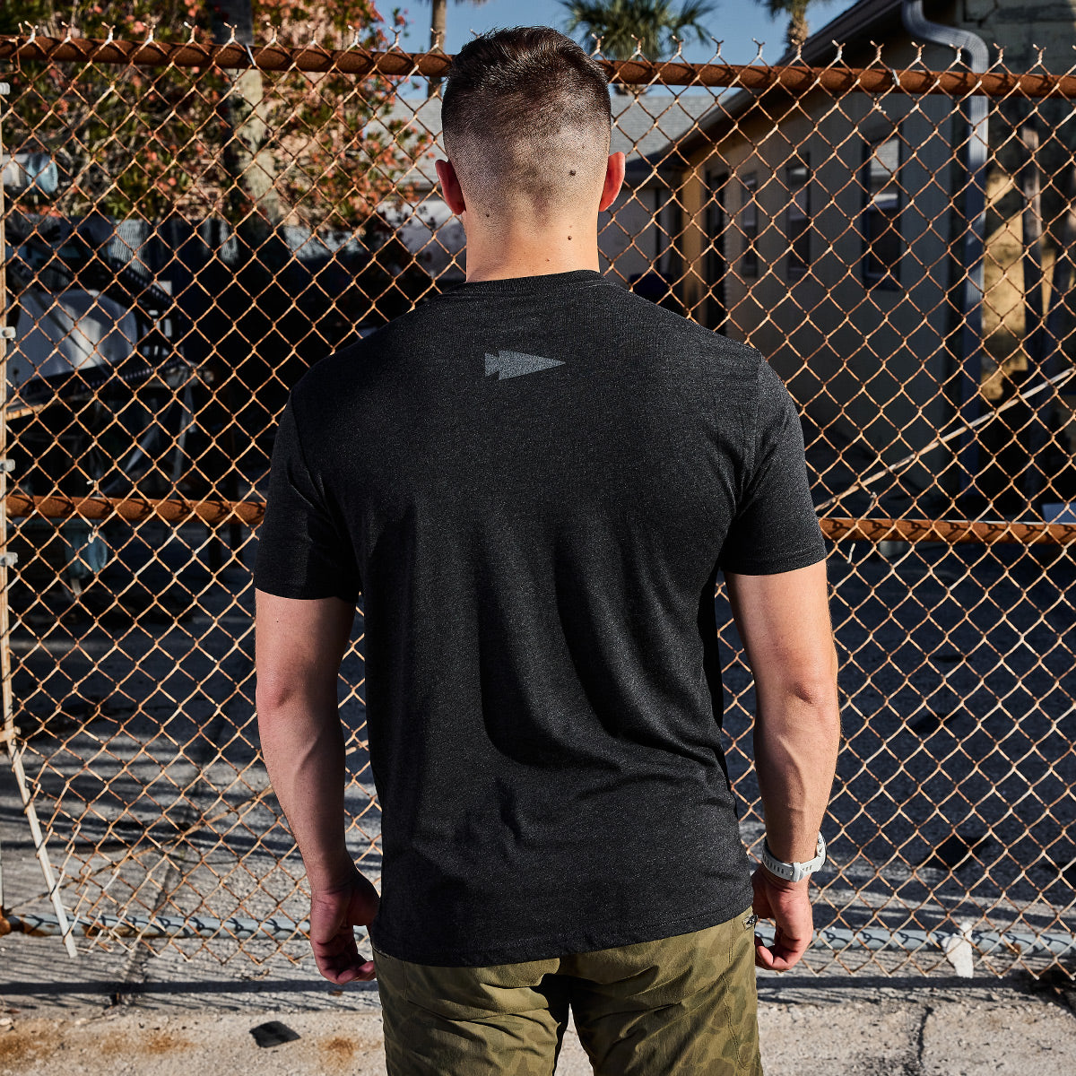 Turning away from the camera, a person displays the GORUCK Spearhead Tee - Tri-Blend in sleek black, paired with olive green pants. They are positioned in front of a chain-link fence under the bright sun, with houses and trees extending into the distance.