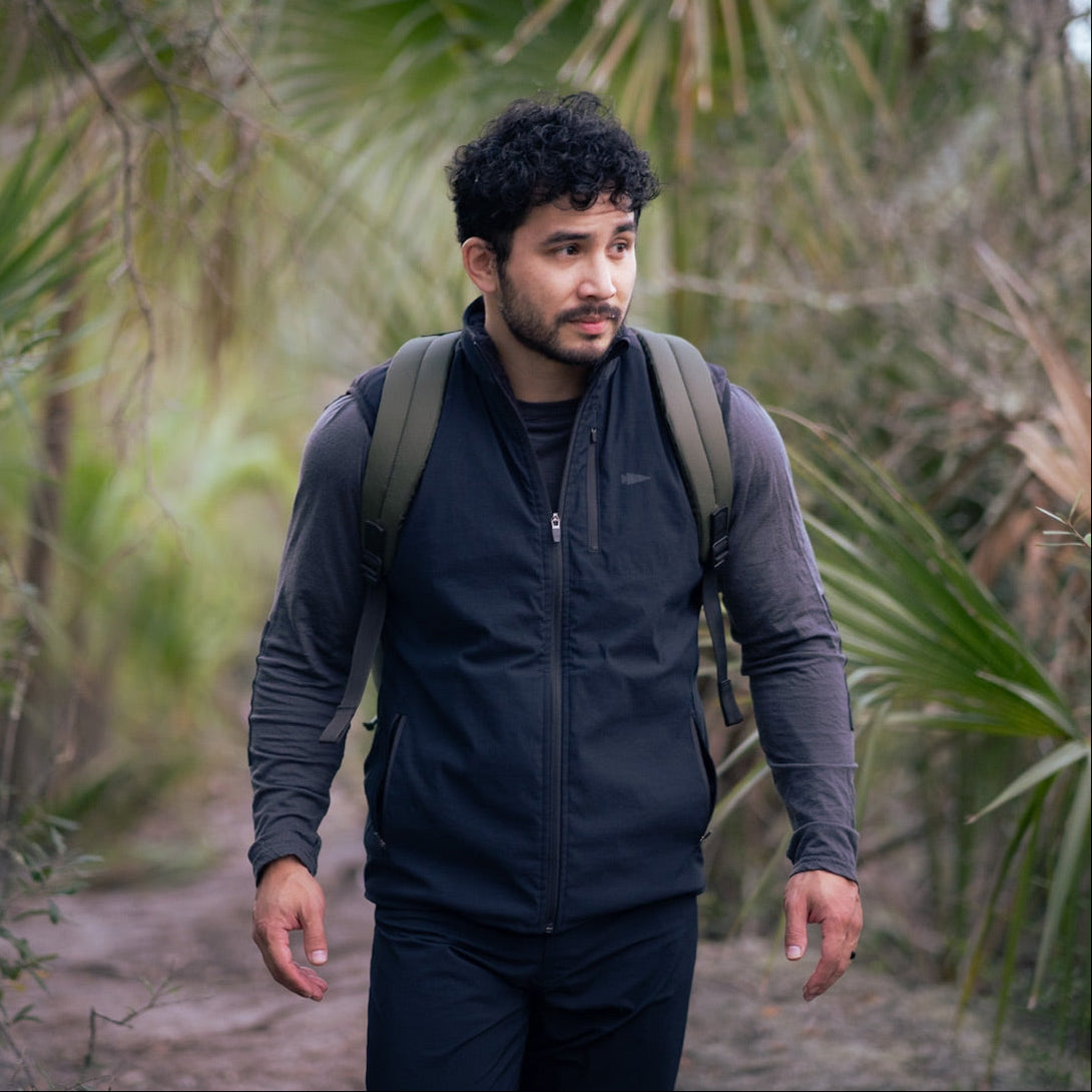 A man with a GORUCK Vest of Power - ToughDry® + Fleece strolls through a forest trail, surrounded by lush greenery.