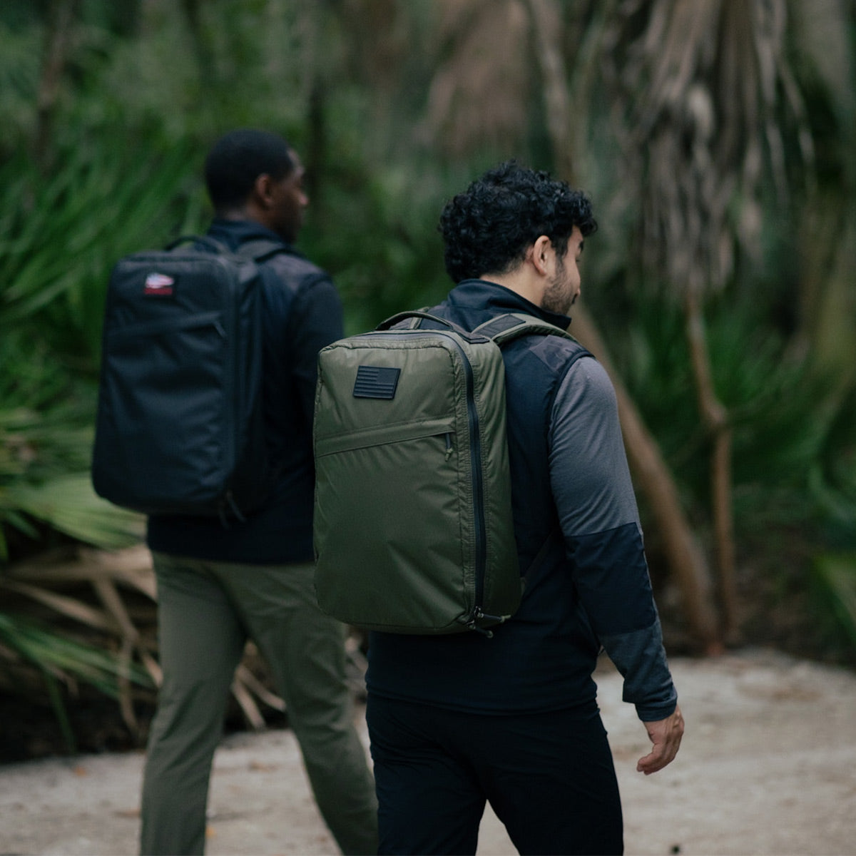 Two people walking through a wooded area with backpacks, one in green and the other in black, each proudly sporting GORUCK's environmentally conscious Vest of Power - ToughDry® + Fleece gear.