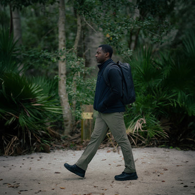 A man, wearing the Vest of Power made from ToughDry® and fleece, carries a GORUCK backpack as he walks along an eco-friendly forest path surrounded by lush greenery.