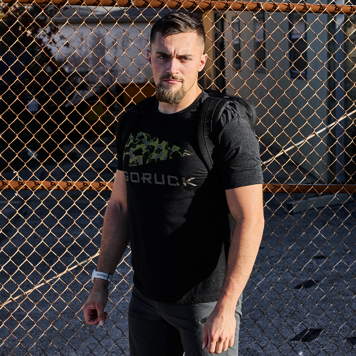 A man with short hair and a beard stands in front of a chain-link fence, embodying the Special Forces roots in his dark vendor-unknown GORUCK Spearhead Tee - Tri-Blend. Sporting a watch and a backpack slung over one shoulder, the sunlight casts shadows on the ground and fence.