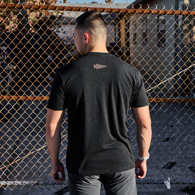 A man with short hair stands facing a chain-link fence, wearing the GORUCK Spearhead Tee - Tri-Blend in black. The T-shirt features a small spearhead logo on the back, and the sun casts intricate shadows from the fence onto the ground.