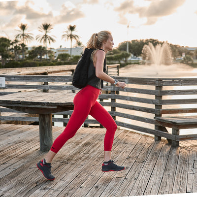 A woman in red leggings and a black tank top, sporting the GORUCK Women's Ballistic Trainers in Wolf Grey and High Risk Red with a Red Reflective Spearhead, jogs along a wooden boardwalk near a lake with a fountain. The sky is partly cloudy, and palm trees sway in the background.