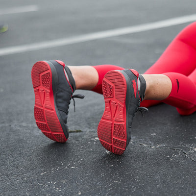 A person wearing the Women's Ballistic Trainers in Wolf Grey and High Risk Red with red reflective spearhead, along with red leggings, is lying on an outdoor surface. The soles of the GORUCK functional fitness footwear are visible, showcasing a textured tread design.