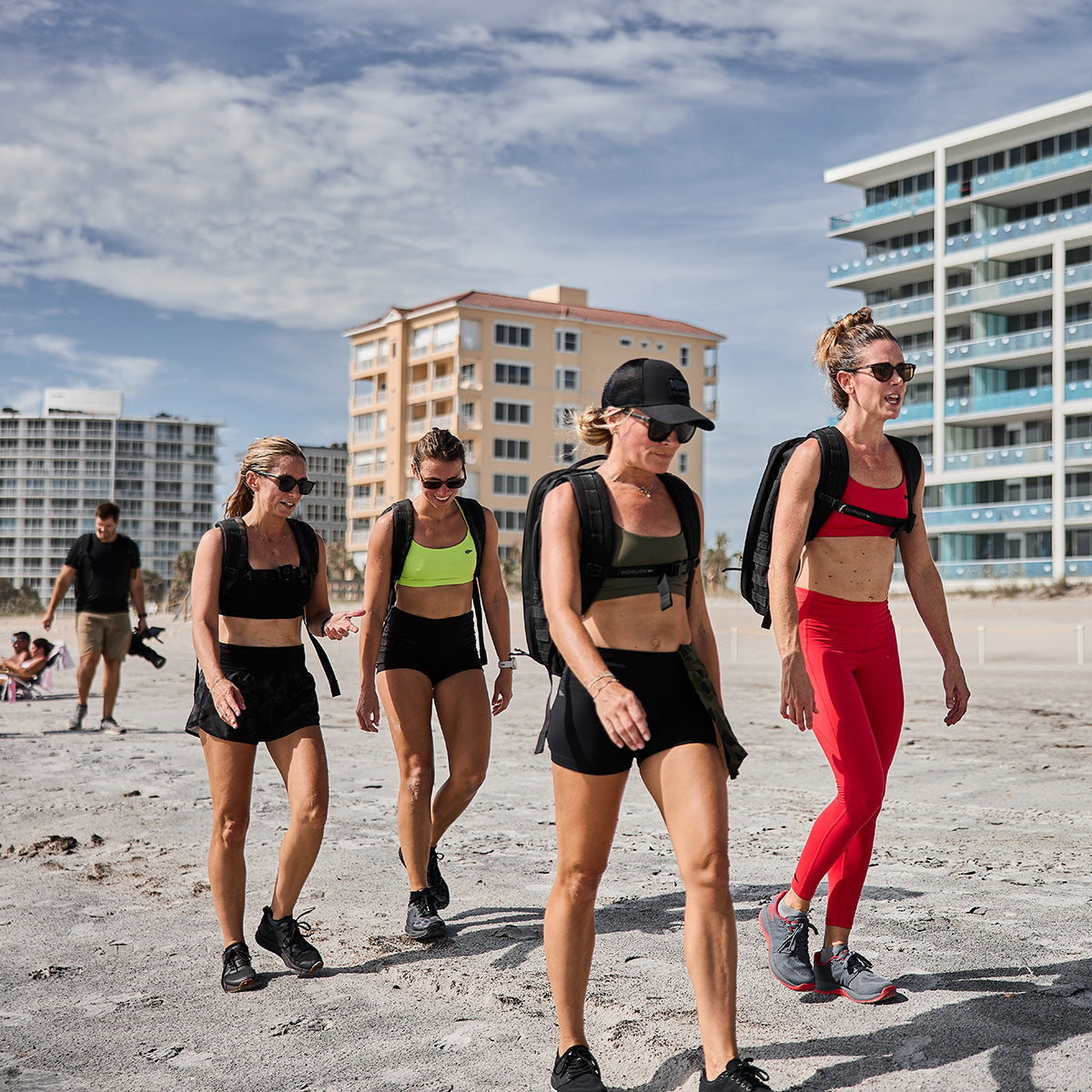 Four women in athletic gear walk along a sunny beach, each carrying a backpack and sporting GORUCK Women's Ballistic Trainers in Wolf Grey with High Risk Red and Red Reflective Spearhead. Behind them are modern beachfront buildings, while a few more people stroll along the shore. The sky is partly cloudy as they benefit from enhanced stability during their walk.