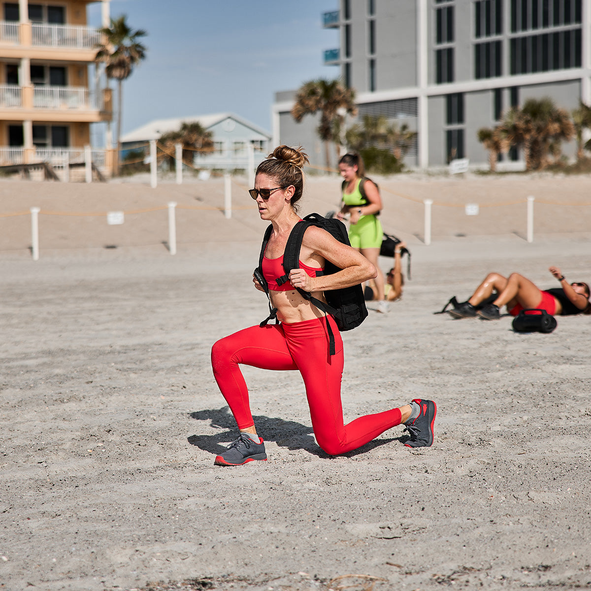 A woman dressed in a red sports outfit performs lunges on a sandy beach while wearing a backpack and showcasing her GORUCK Women's Ballistic Trainers in Wolf Grey and High Risk Red. Other people can be seen near buildings, enjoying the sunny day with palm trees nearby.