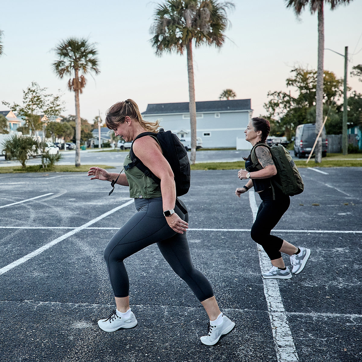 Two women, each equipped with GORUCK Women's Ballistic Trainers in Lunar Rock and Charcoal featuring a Silver Reflective Spearhead, dash through an empty parking lot. In the sunlight, palm trees and houses set the backdrop for their run. One wears gray leggings while the other chooses black, both benefiting from 3X Support™ for enhanced comfort and durability on their journey.