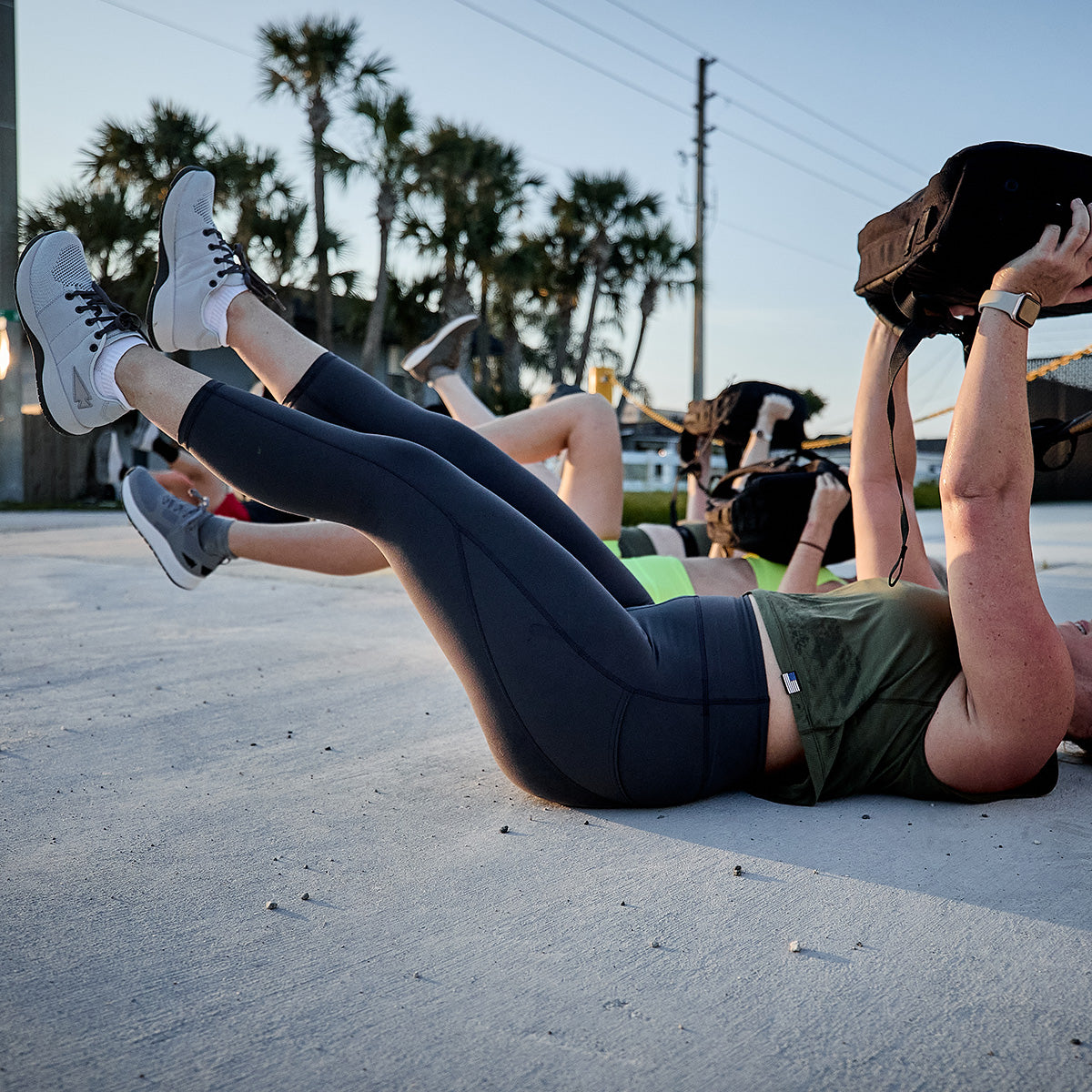 Two individuals exercise on a concrete surface under clear skies, effortlessly raising their legs while holding weights. They're wearing athletic gear and the Women's Ballistic Trainers in Lunar Rock + Charcoal with Silver Reflective Spearhead by GORUCK, featuring robust CORDURA® Ballistic Nylon. The scene is beautifully set against palm trees swaying gently in the background.