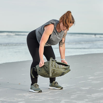 On the beach, a dedicated fitness enthusiast in athletic wear tackles a Women's Rough Runner - Earth sandbag workout. With ocean waves crashing, their routine displays resilience against the elements.