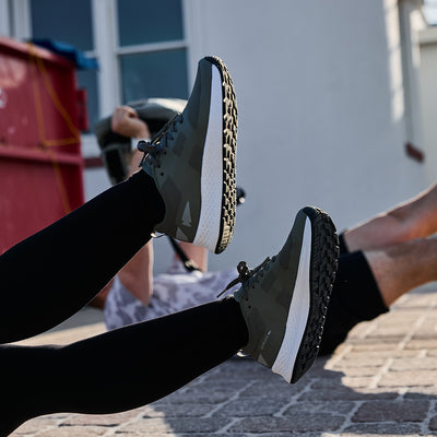 Two people engage in outdoor exercise, showcasing the Women's Rough Runner - Earth shoes as they raise their legs on a stone surface. These high-mileage shoes feature Gradient Density cushioning for superior comfort and support.