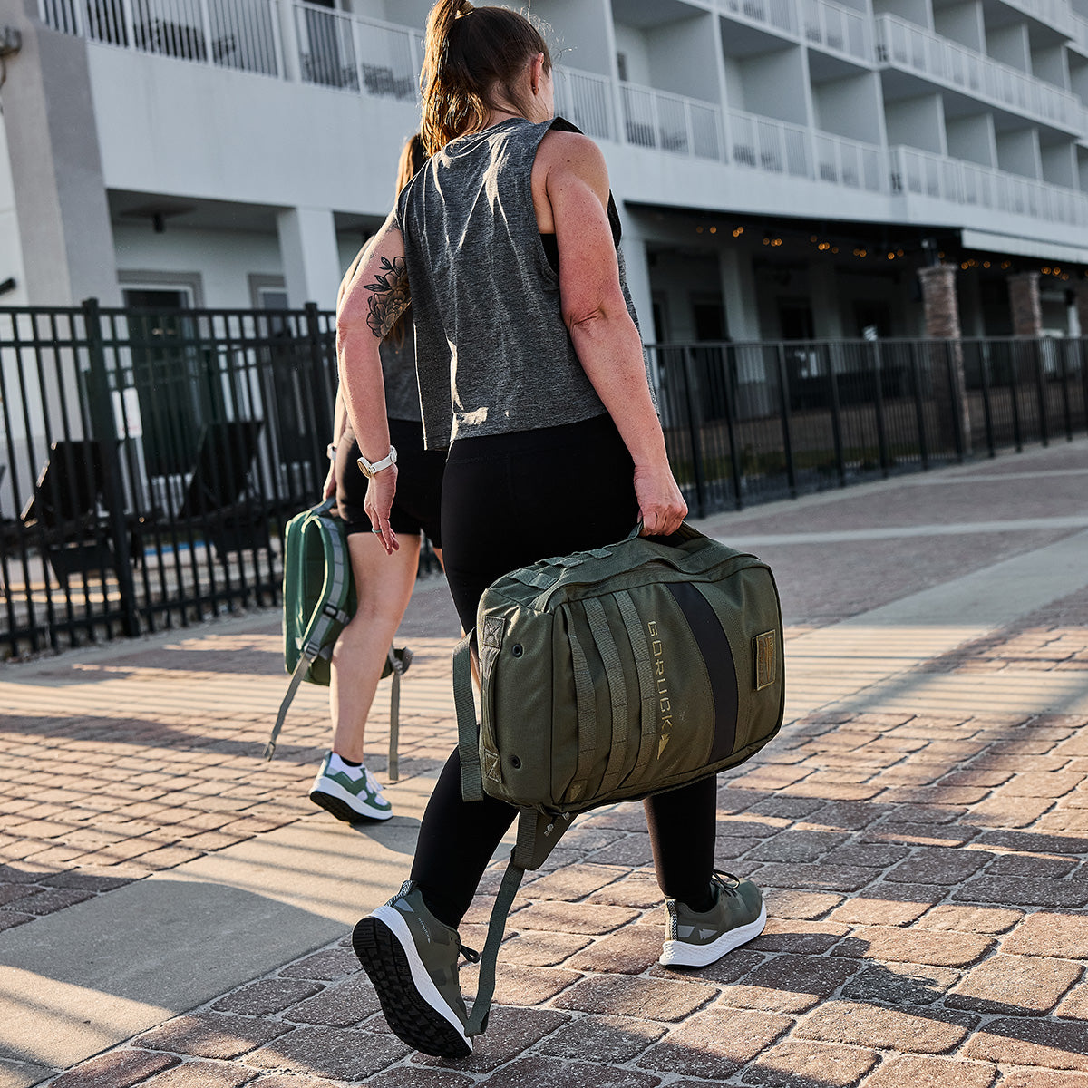 Two women walk outdoors with their Women's Rough Runner - Earth duffle bags near a building with balconies, ready for the challenge.