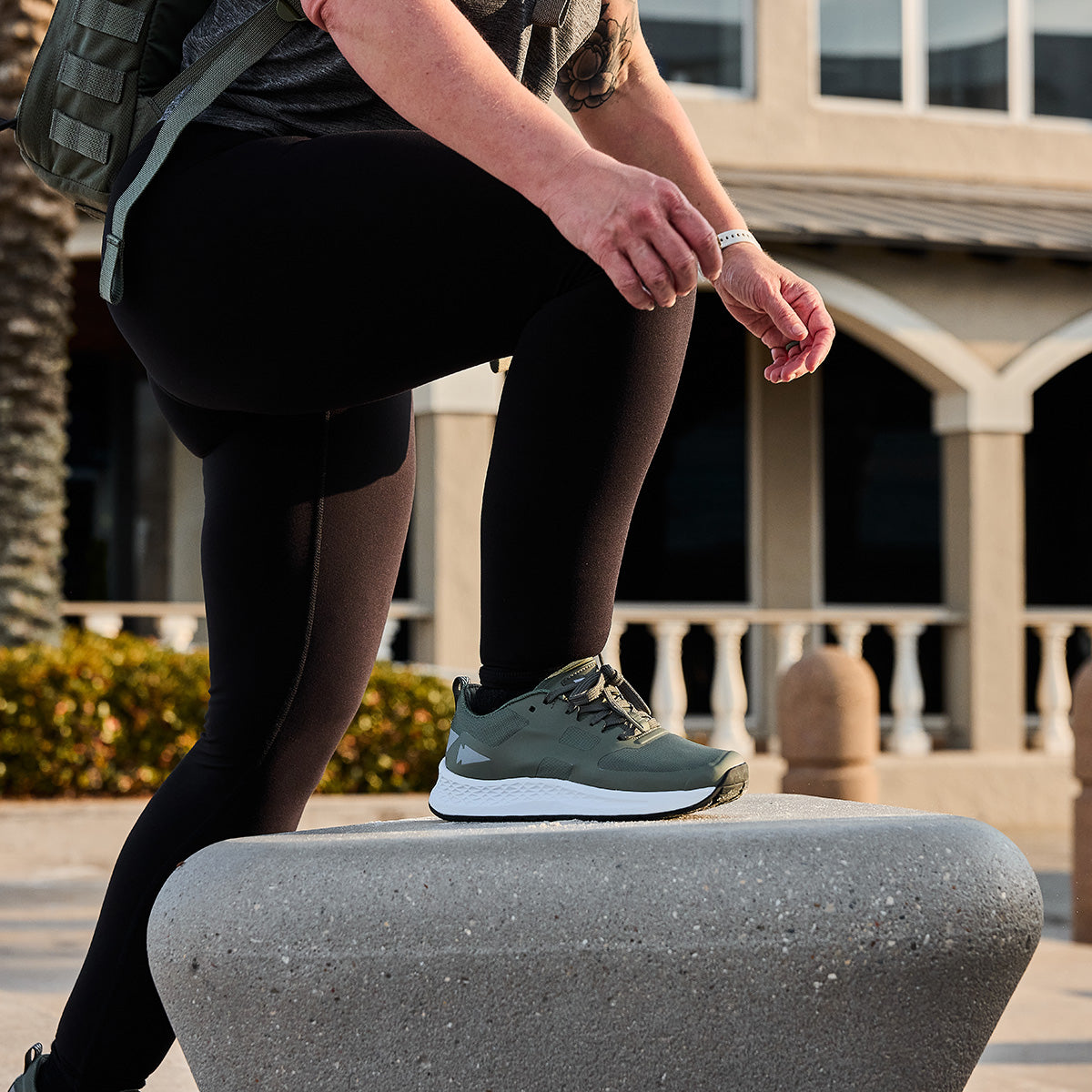 A woman confidently steps on the rugged outdoor concrete near a building, embodying the spirit of the Women's Rough Runner - Earth in her green sneakers.