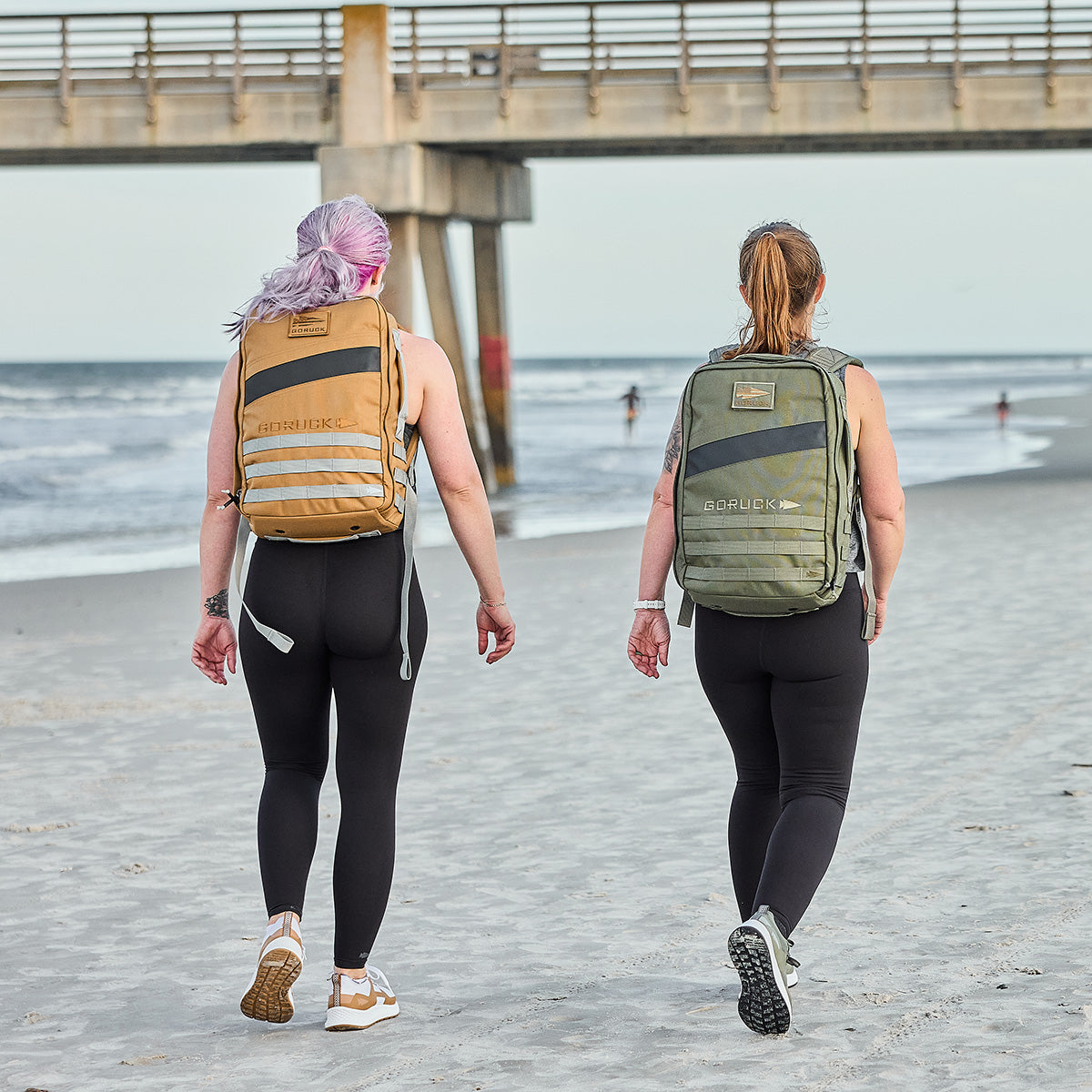 Two women in Women's Rough Runner - Earth backpacks traverse a beach by the pier, dressed in athletic wear and ready for high-mileage adventures.