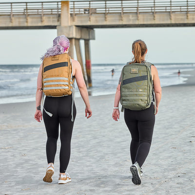 Two women in Women's Rough Runner - Earth backpacks traverse a beach by the pier, dressed in athletic wear and ready for high-mileage adventures.