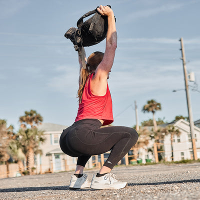 Sporting the Women's Rough Runner in light and dark grey, she squats outdoors, lifting a backpack overhead, embodying its adventurous spirit.