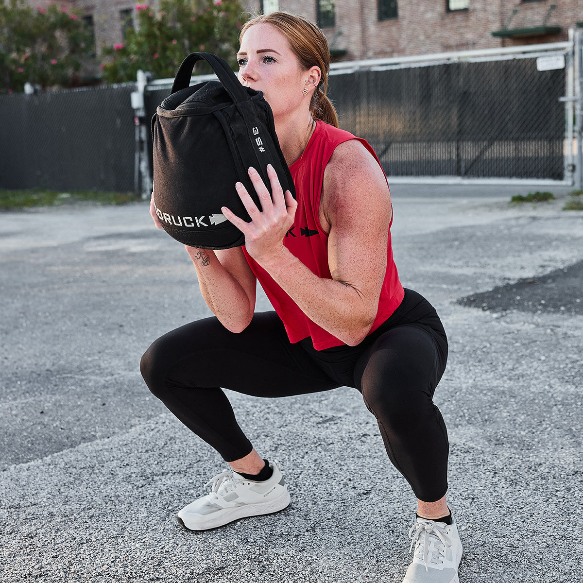 A woman in a red top and black leggings performs a squat outdoors with a black sandbag, resembling the Women's Rough Runner - Light Grey + Dark Grey, exhibiting strength and stability like an EVA Midsole.