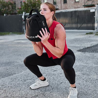 A woman in a red top and black leggings performs a squat outdoors with a black sandbag, resembling the Women's Rough Runner - Light Grey + Dark Grey, exhibiting strength and stability like an EVA Midsole.