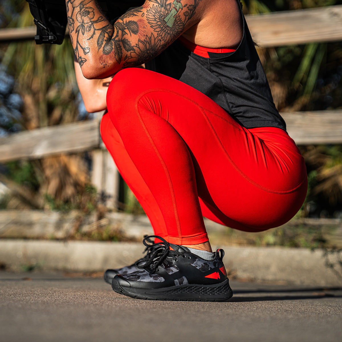 On a sunny day, a person crouches outdoors, wearing the Women's Rough Runner in Midnight Frogskin + High Risk Red from GORUCK. Their red leggings match their black top and shoes perfectly, while their tattoo-covered arm stands out against the wooden fence and lush greenery behind them.