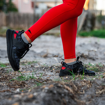 A close-up shows someone walking outdoors in the Women's Rough Runner - Midnight Frogskin + High Risk Red from GORUCK, featuring bright red leggings and black athletic shoes with red accents. The rugged grassy terrain hints at an off-road adventure, ideal for enjoying the EVA Midsole's optimal comfort and support with every stride.