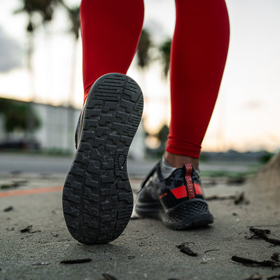 A person clad in high risk red leggings strolls down the sidewalk, highlighting the EVA midsole of their GORUCK black athletic shoe with a high risk red accent from the Women's Rough Runner - Midnight Frogskin collection. The backdrop is a blur of bustling street life framed by palm trees.