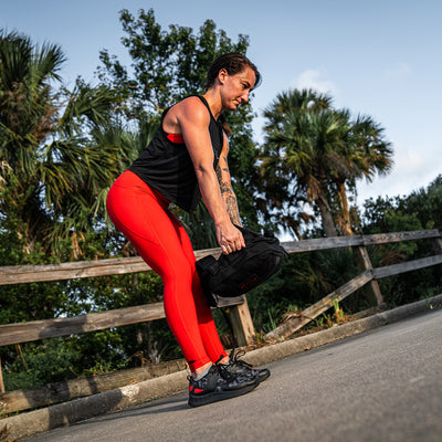 Decked out in GORUCK's Women's Rough Runner - Midnight Frogskin + High Risk Red, a person effortlessly lifts a black weighted bag outdoors. They embody the spirit of a true Rough Runner on a paved path, surrounded by lush green trees and wooden fences under a clear sky.