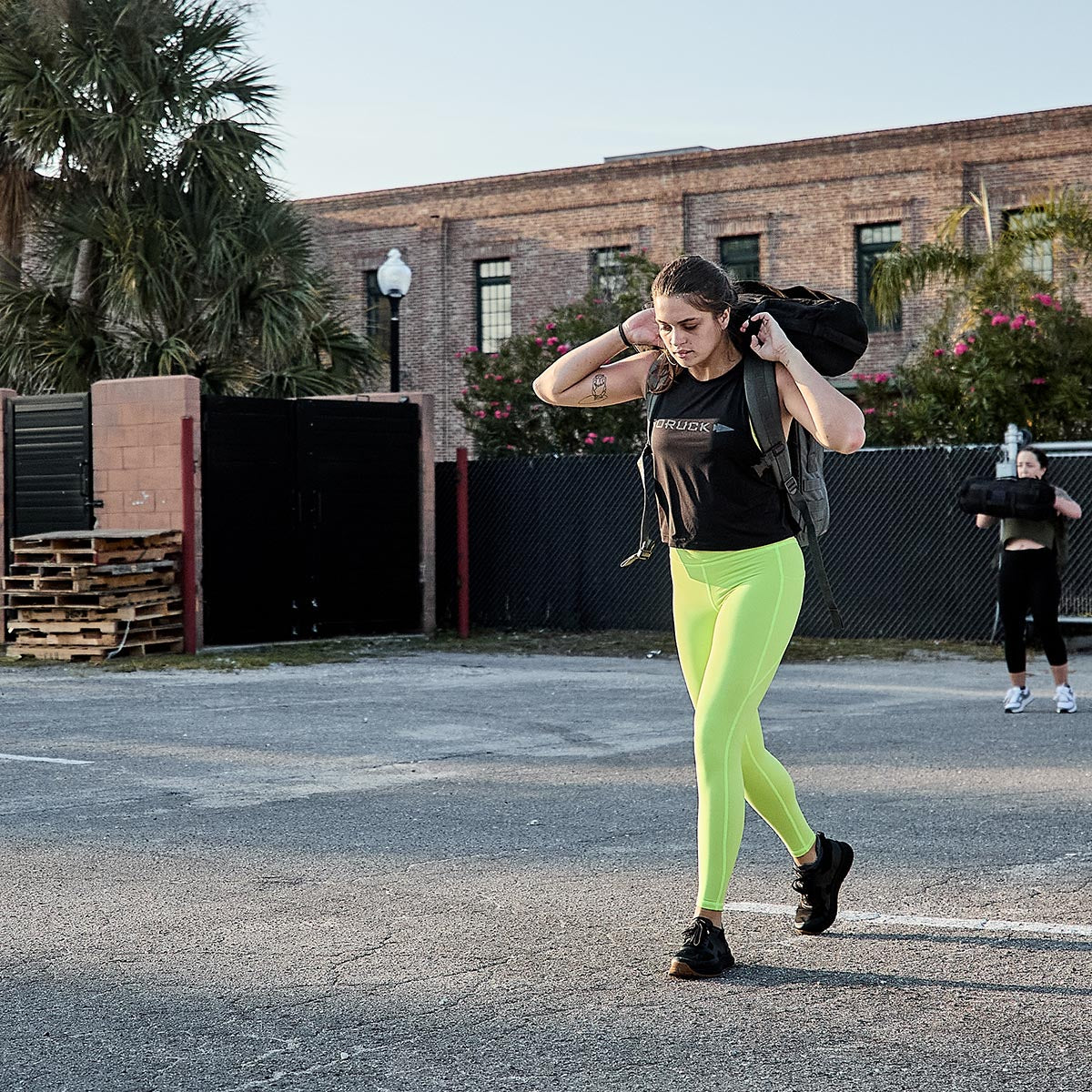 A woman wearing GORUCK's Women’s Training Leggings Pocket - ToughFlex in bright yellow and a black top strolls through a parking area, holding a black bag over her shoulder. Meanwhile, another person stretches nearby. A brick building and palm trees provide the backdrop to this vibrant scene.