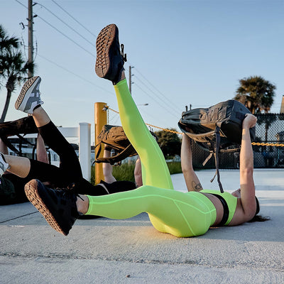 Two individuals exercise outdoors on a sunny day, holding weighted bags while lying on their backs. One is wearing bright yellow athletic wear, and the other sports GORUCK Women’s Training Leggings Pocket - ToughFlex in black. Both have one leg raised in the air, with palm trees swaying gently in the background.