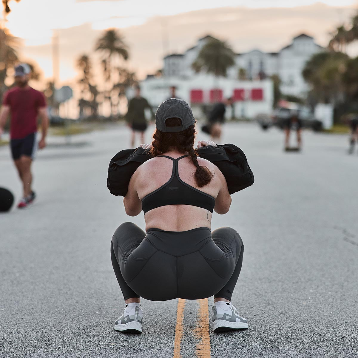 A person wearing a sports bra, GORUCK Women’s Training Leggings Pocket - ToughFlex, and a cap performs a squat with a weighted sandbag on a street. Other people and palm trees are in the background, with soft sunset lighting setting the scene.