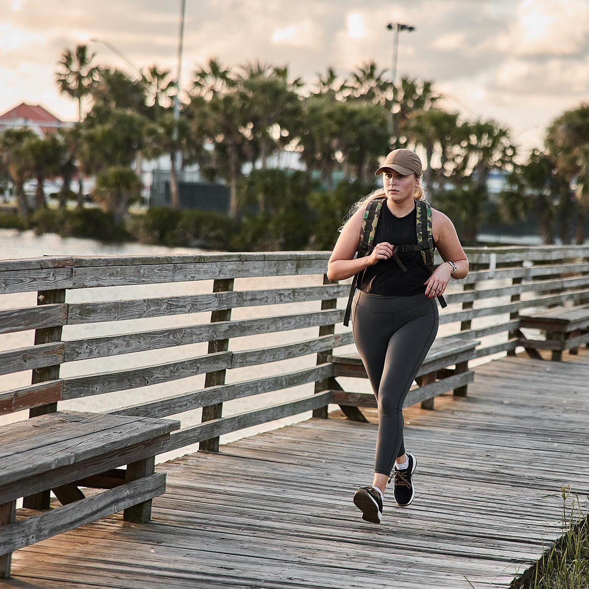 A person strolls along a wooden boardwalk near tranquil waters, surrounded by palm trees. Dressed in athletic attire with GORUCK’s Women’s Training Leggings Pocket - ToughFlex and a cap, they carry a backpack. The sky is cloudy, creating a calm and serene atmosphere.