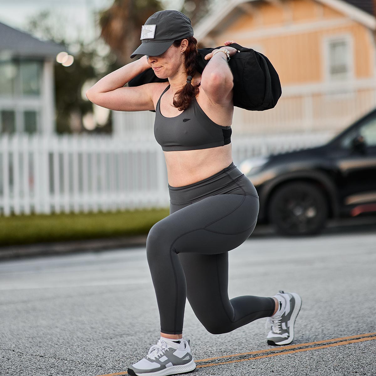 A woman in athletic wear performs a lunge exercise on a residential street, showcasing her GORUCK Women’s Training Leggings Pocket - ToughFlex and holding a black bag over her shoulder. She wears a cap and sneakers, with houses and a car visible in the background.