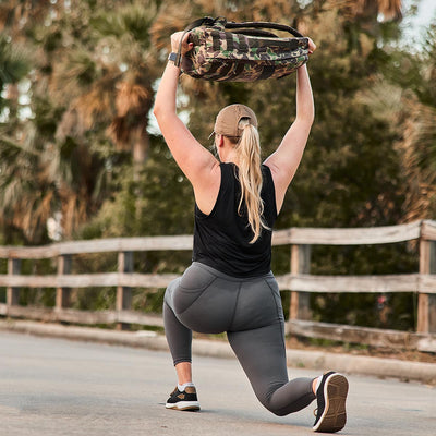 The person, sporting GORUCK’s Women's Training Leggings Pocket - ToughFlex and a camo-patterned weight bag lifted overhead, lunges gracefully along an outdoor path lined with trees and wooden fences. This scene embodies focus and strength against the backdrop of nature's tranquility.