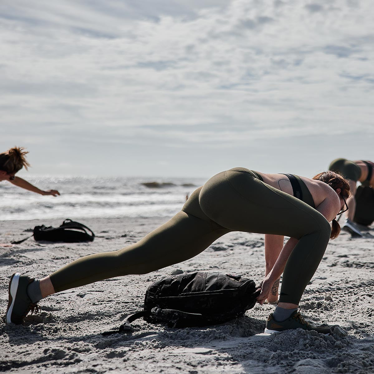 A woman stretches on a sandy beach as others linger in the background beneath an overcast sky. She leans forward, touching the ground with one hand while wearing GORUCK Women’s Training Leggings Pocket crafted from ToughFlex fabric, with a bag placed nearby.