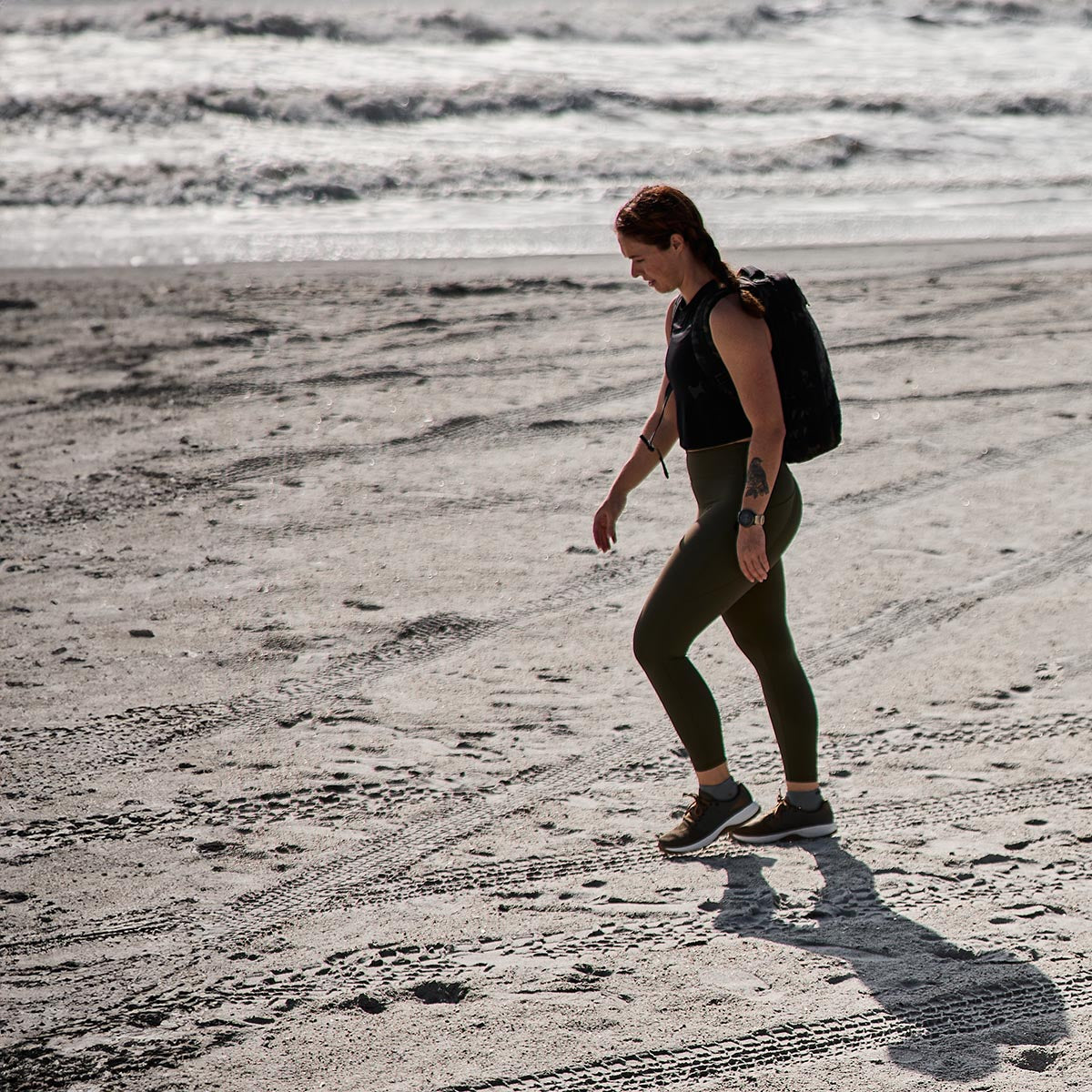 A person with long hair, sporting high-waisted GORUCK Women's Training Leggings Pocket - ToughFlex and carrying a backpack, strolls along a sandy beach. The ocean waves are visible in the background, with sunlight casting shadows on the sand.