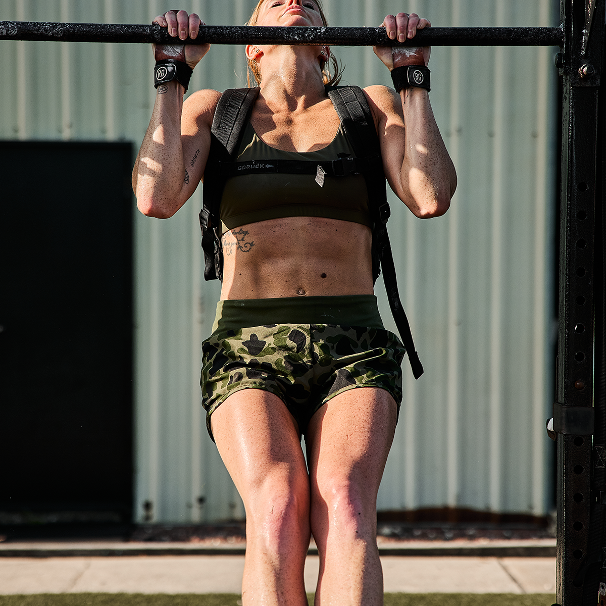 A woman in GORUCK’s ToughStretch Women's Training Shorts and a weighted vest performs a pull-up on an outdoor bar. Her focused effort is evident as the sun highlights her muscular physique, casting shadows. A building with a metal wall stands in the background, adding an urban vibe to her intense workout session.