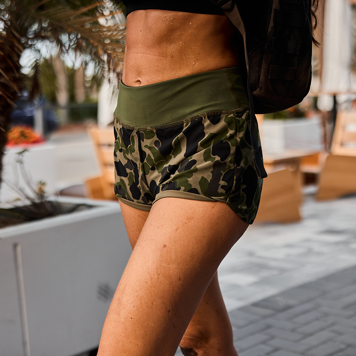 A person walks outdoors, wearing GORUCK's Women’s Training Shorts - ToughStretch and carrying a backpack. The emphasis is on their lower body, showcasing the squat-proof durability of the shorts. In the background, plants and outdoor seating enjoy the sun's warm glow.