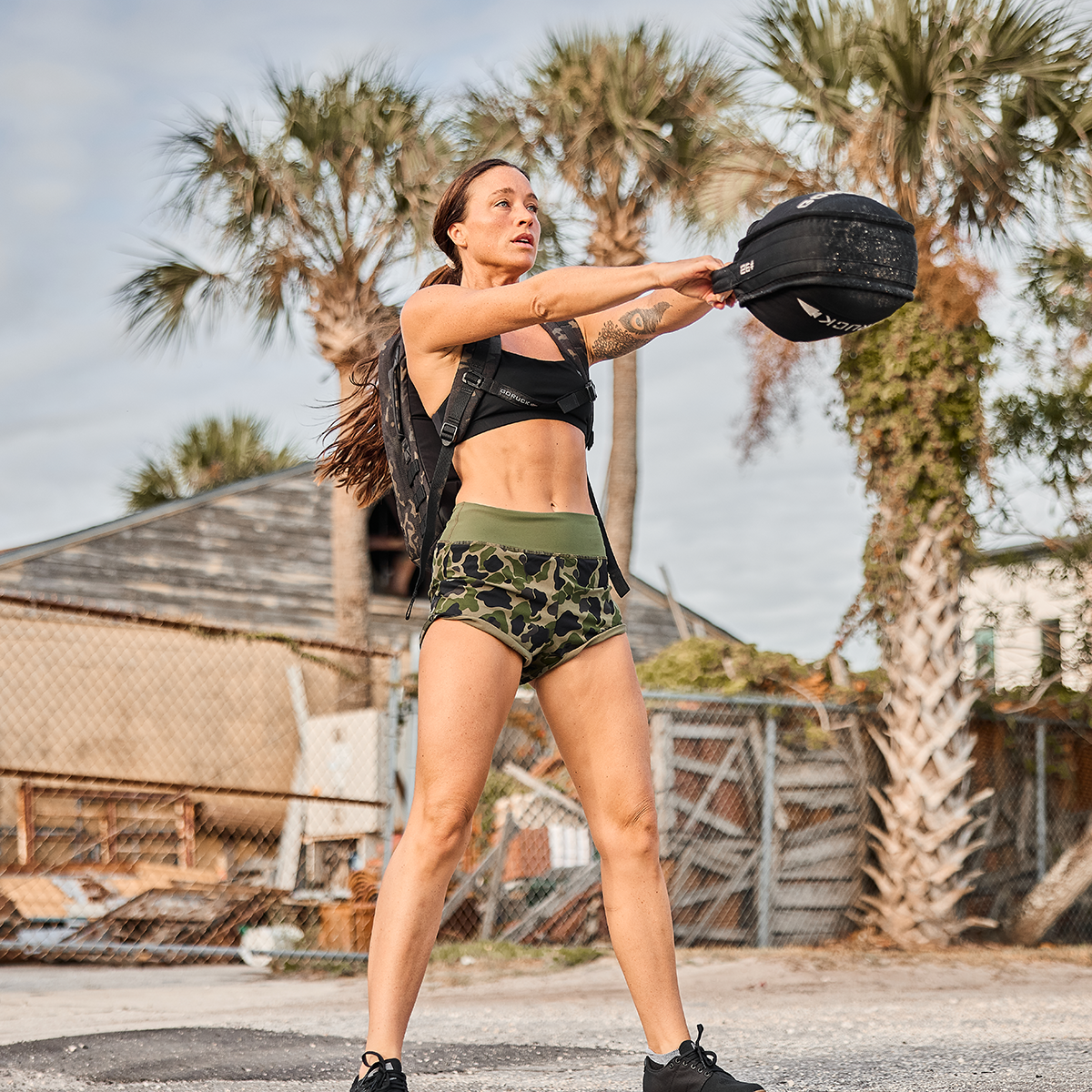 A woman in workout attire is lifting a sandbag during an outdoor exercise, showcasing her squat-proof movement. She is wearing a black sports bra, GORUCK Women's Training Shorts in ToughStretch camouflage fabric, and boots. Palm trees and a wooden building are in the background under a partly cloudy sky.