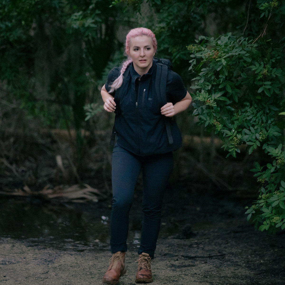 A person with pink hair hikes on a forest trail in a black GORUCK outfit and brown boots. Their Women's Vest of Power - ToughDry® + Fleece fits snugly as they enjoy the lush greenery.