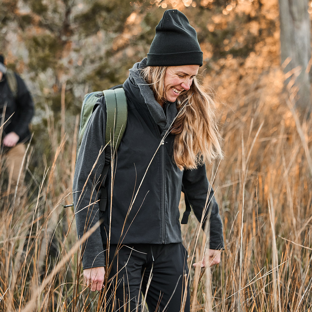 Smiling individual walks through tall grass, wearing a black beanie and the Women's Vest of Power featuring ToughDry® + Fleece, carrying a GORUCK backpack.