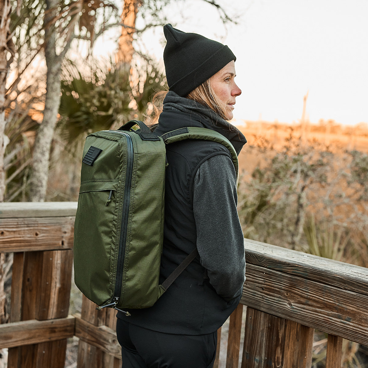 A person wearing the Women's Vest of Power - ToughDry® + Fleece, black attire, and a beanie stands confidently on a wooden path in nature with a green GORUCK backpack.