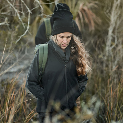 A woman in a Women's Vest of Power - ToughDry® + Fleece, embodying GORUCK's rugged style, strolls through tall grass. Another figure lingers in the background, enhancing the scene's adventurous spirit.