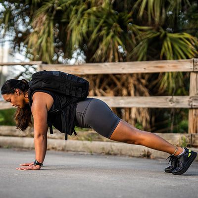 A woman in athletic wear performs a plank exercise on a paved surface, demonstrating the stay-put technology of her GORUCK Women’s Biker Shorts - ToughFlex. She is wearing a backpack and sneakers. In the background, there is a wooden fence and palm trees.