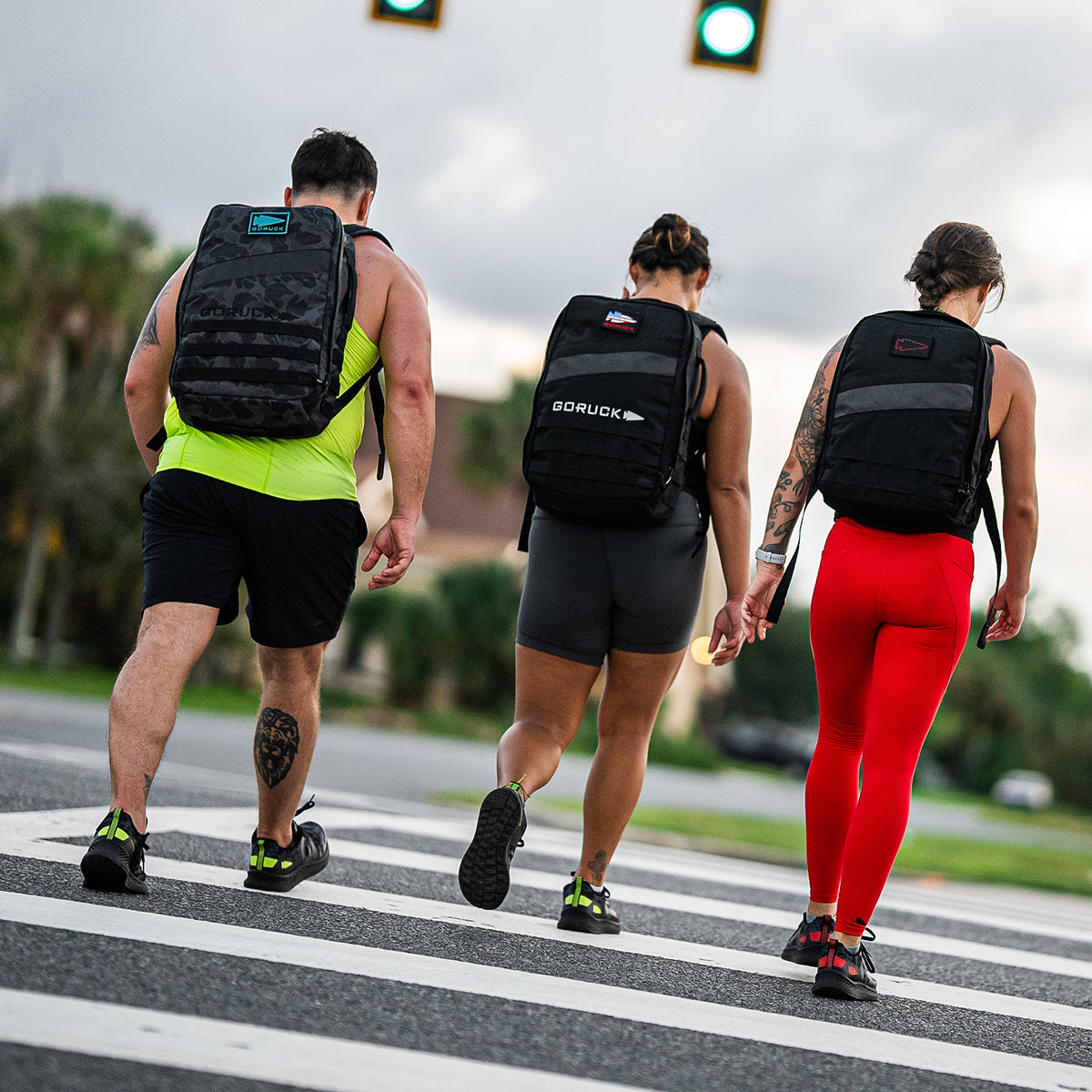 Sporting Women's Rough Runner - Midnight Frogskin + Acid Lime by GORUCK, three individuals with backpacks cross a street at a crosswalk, clad in lightweight running shoes and athletic clothing such as a tank top and bright red leggings. The scene is further energized by traffic lights and a cloudy sky, evoking the spirit of a Rough Runner challenge.