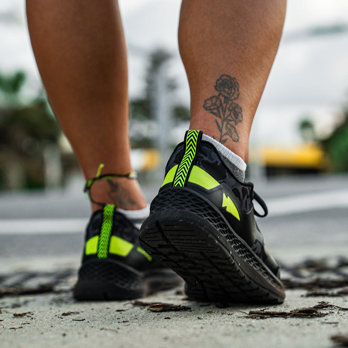 Close-up of a person walking on a street in GORUCK's Women's Rough Runner - Midnight Frogskin + Acid Lime shoes, featuring bright yellow accents and a gradient density EVA midsole. The person's ankle showcases a rose tattoo, with an overcast sky and greenery blurred in the background, embodying the spirit of a Rough Runner journey.